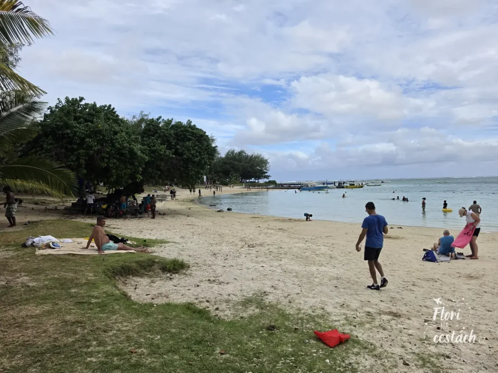 eřejná písečná pláž Blue Bay beach na Mauriciu s odpočívajícími lidmi, zelenými stromy a loděmi kotvícími v mělké zátoce